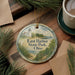 A decorative ornament featuring a lighthouse and a scenic landscape, with the text "East Harbor State Park, Ohio" printed on it. The ornament is placed on a wooden surface, surrounded by pine branches and a cup of coffee.