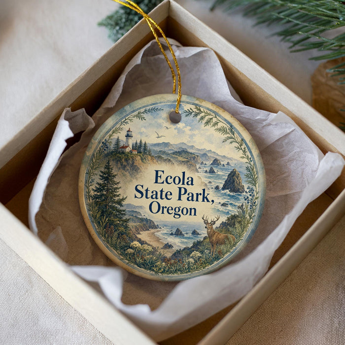 A decorative ornament in a box, featuring a scenic landscape with mountains, trees, and a lighthouse, with the text "Ecola State Park, Oregon" printed on it.