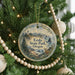 A round ornament hanging on a Christmas tree, featuring a scenic landscape with mountains, a lighthouse, and a body of water.