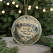 A round ornament with a scenic landscape of Oregon, featuring a lighthouse, mountains, and a deer, is displayed in front of a blurred background with Christmas lights.