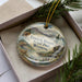 A decorative ornament in a box, featuring a scenic landscape with mountains, waterfalls, and wildlife.