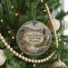 A round ornament hanging on a Christmas tree, featuring a scenic landscape with mountains, a waterfall, and a deer.