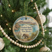 A circular ornament hanging on a Christmas tree, featuring an illustration of an elephant and a scenic landscape with mountains, water, and a boat.