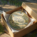 A wooden box containing a round ornament with a scenic landscape image, including a body of water, mountains, and a small building.