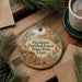 A decorative ornament with a forest scene and text, placed on a wooden surface next to a cup of coffee and pine branches.