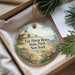 A decorative ornament with a lighthouse and beach scene, placed in a box with pine branches.