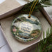 A decorative ornament in a box, featuring a scenic landscape with a bridge, trees, and mountains.