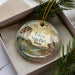 A decorative ornament in a box, featuring a scenic landscape with a bird perched on a rock, surrounded by greenery.