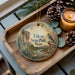 A decorative ornament with a bird and a scenic landscape, featuring the text "Falcon State Park, Texas" on it, is displayed on a wooden tray alongside pine cones and a lit candle.