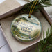 A decorative ornament in a box, featuring a scenic landscape with a body of water, trees, and a bird.