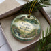 A decorative ornament in a box, featuring a scenic landscape with a building, trees, and a body of water.