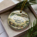 A decorative ornament in a box, featuring an illustration of a historic house surrounded by trees and flowers.