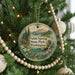 A circular ornament hanging on a Christmas tree, featuring a scenic landscape with mountains, a lake, and a cabin.