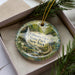 A decorative ornament in a box, featuring a scenic landscape with a waterfall and greenery.