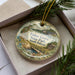 A decorative ornament in a box, featuring a scenic landscape with a bridge, trees, and a cabin, with the text "Fisher Grove State Park, South Dakota" printed on it.