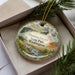 A decorative ornament in a box, featuring a scenic landscape with a river, mountains, and pine trees.