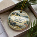 A decorative ornament in a box, featuring a scenic landscape with a house, trees, and a cannon, along with the text "Fort Columbia State Park, Washington".