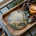 A decorative ornament featuring a lighthouse and a coastal scene, placed on a wooden tray alongside pine cones and a lit candle.