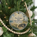 A round ornament hanging on a Christmas tree, featuring a lighthouse and a coastal scene.