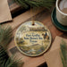 A round ornament with a picture of Fort Griffin, a historic site in Texas, is displayed on a wooden surface alongside a cup of coffee and some pine branches.