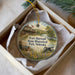 A round ornament with a picture of a historical building and a wagon, surrounded by flowers, placed in a box with a white tissue paper lining.