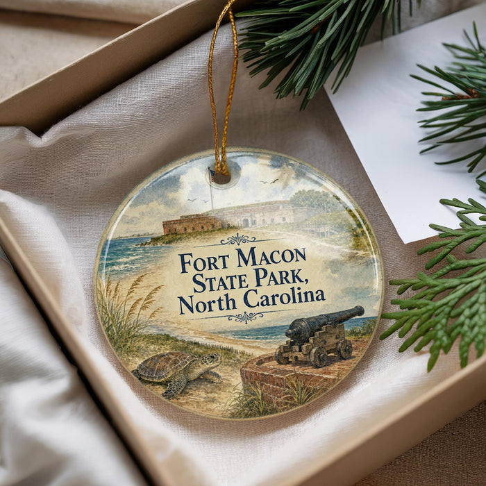 A round ornament with a picture of Fort Macon State Park, North Carolina, featuring a cannon and a turtle, is displayed in a box with pine branches.