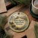 A decorative ornament with a scenic image of Fort Owen State Park, Montana, hanging on a wooden surface.