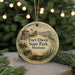 A round ornament with a painting of Fort Owen State Park in Montana, featuring a wagon and a building, is displayed on a Christmas tree.