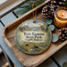 A decorative ornament with a scenic image of Fort Ransom State Park, North Dakota, featuring a house, a river, and wildlife. The ornament is placed on a wooden tray, with pine cones and a lit candle nearby.