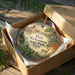 A round ornament in a box, featuring a scenic landscape with a castle, trees, and a deer, along with the text "Fort Richardson State Park, Texas".
