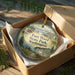 A round ornament in a box, featuring a scenic landscape with a house and trees, with the text "Fort Washington State Park, Washington" printed on it.