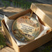 a circular ornament with a scenic landscape, placed inside a brown cardboard box on a wooden surface.