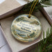 A decorative ornament in a box, featuring a scenic landscape with a lake, mountains, and a deer.