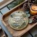 A decorative ornament with a scenic lake landscape and pine cones sits on a wooden tray, accompanied by a lit candle and pine cones.