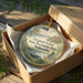 A round ornament in a box, featuring a scenic landscape with a lake, mountains, and a deer, along with the text "Fremont Lakes State Recreation Area, Nebraska".