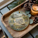 A decorative ornament featuring a hotel and a scenic landscape, placed on a wooden tray alongside pine cones and a lit candle.