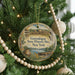 A round ornament hanging on a Christmas tree, featuring a scenic landscape with a river, a building, and a boat.