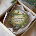 A round ornament with a scenic landscape of a lake, mountains, and a bird in flight, placed in a box with a white tissue paper lining.