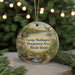 A round ornament with a scenic landscape featuring a lake, mountains, and wildlife, including a bird in flight, hangs in front of a blurred background of Christmas lights.