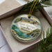 A decorative ornament in a box, featuring a scenic landscape with a river, mountains, and a house, with the text "Giant Springs State Park, Montana" printed on it.