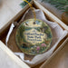 A decorative ornament in a box, featuring a scenic landscape with a river, trees, and wildlife, including a deer and a bird.