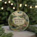 A round ornament with a scenic landscape of a river, bridge, and wildlife, including a deer, is displayed in front of a blurred background of Christmas lights.