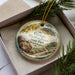 A decorative ornament with a scenic image of the Ginkgo Petrified Forest in Washington State, Washington, placed in a box with pine branches.