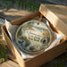 A round ornament in a box, featuring a scenic landscape with a lake, trees, and a house, with the text "Glimmerglass State Park, New York" printed on it.