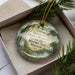 A decorative ornament with a landscape scene and text, placed in a box with pine branches.