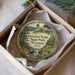 A decorative ornament in a box, featuring a scenic landscape with a waterfall and a cabin, with the text "Governor Dodge State Park, Wisconsin" printed on it.