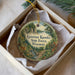 A round ornament with a scenic forest landscape and a deer, placed in a box with a white tissue paper lining.