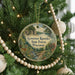 A round ornament hanging on a Christmas tree, featuring a scenic image of a forest and a deer.