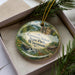 A decorative ornament in a box, featuring a scenic landscape with a waterfall and a cabin, along with the text "Governor Nelson State Park, Wisconsin".