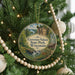 A circular ornament hanging on a Christmas tree, featuring a scenic landscape with a waterfall and a cabin in the background.